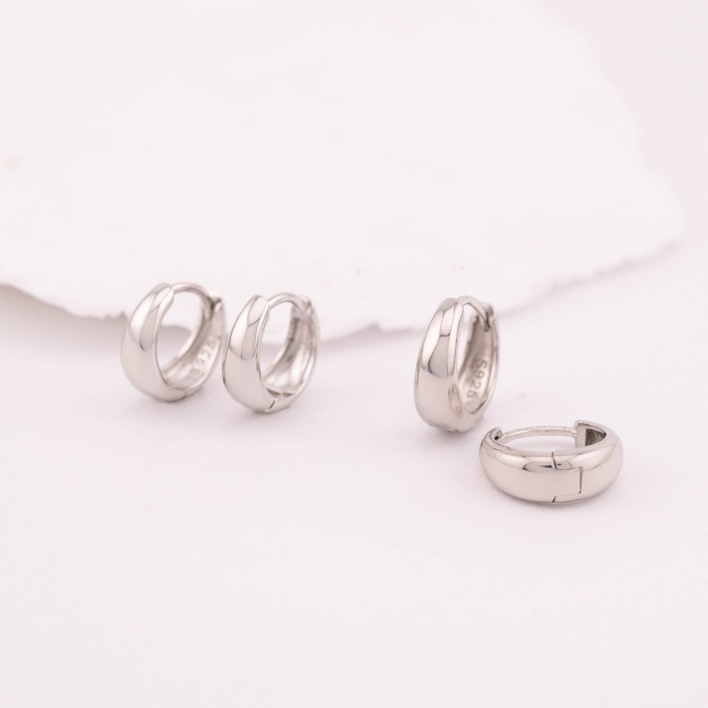 Top front view of two sterling silver huggie earrings on a white surface, showcasing smooth, rounded shapes.